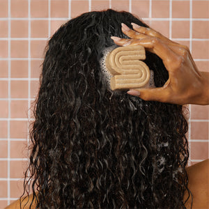 Female model with damp curly hair using a sudsy Kitsch coconut oil shampoo bar on their hair with a pink tile background