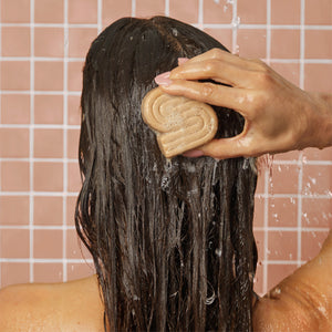 Female model with damp straight hair using a sudsy Kitsch coconut oil shampoo bar on their hair with a pink tile background