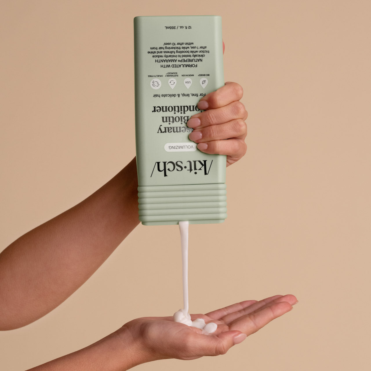 Hand holding a green bottle of Kitsch conditioner with white conditioner being dispensed onto another hand against a beige background.