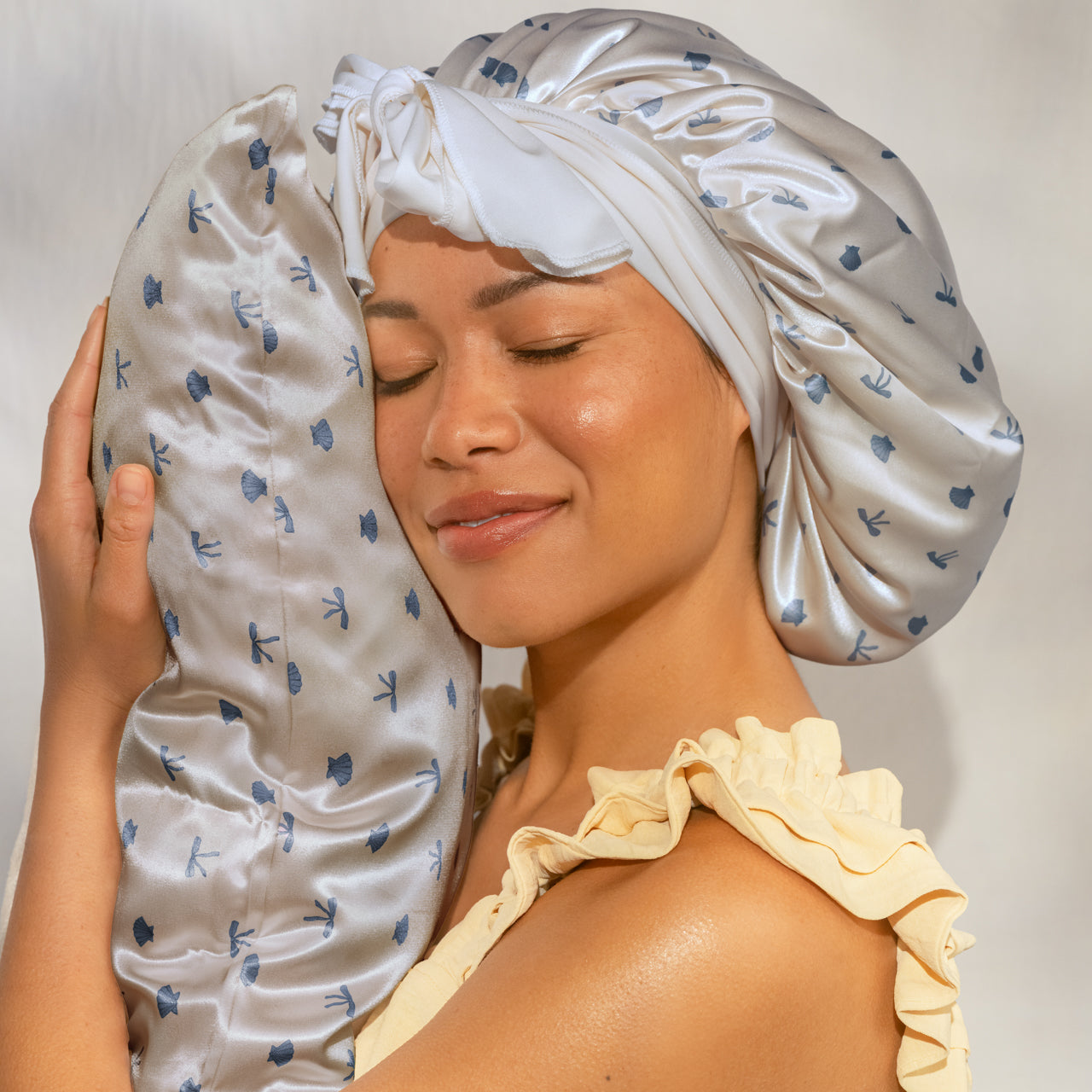 Female model in yellow ruffled top wearing satin bonnet, cheek resting on KITSCH Satin Pillowcase in Pearlized Cream & Coastal Blue Print — eyes closed, serene beauty lifestyle shot — Reduces friction against hair & skin, tames frizz & reduces breakage — For hair & skin-loving comfort