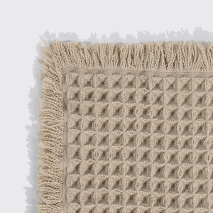Close-up of a camel waffle-knit bath mat's corner, showing the woven texture and fringed trim.