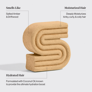 Beige textured shampoo bar with text describing its properties on a light gray background. Properties include: Smells like salted amber & driftwood, moisturized hair - deeply moisturizes kinky, curly, & coily hair, hydrated hair - formulated with coconut oil, known to provide the ultimate hydration boost
