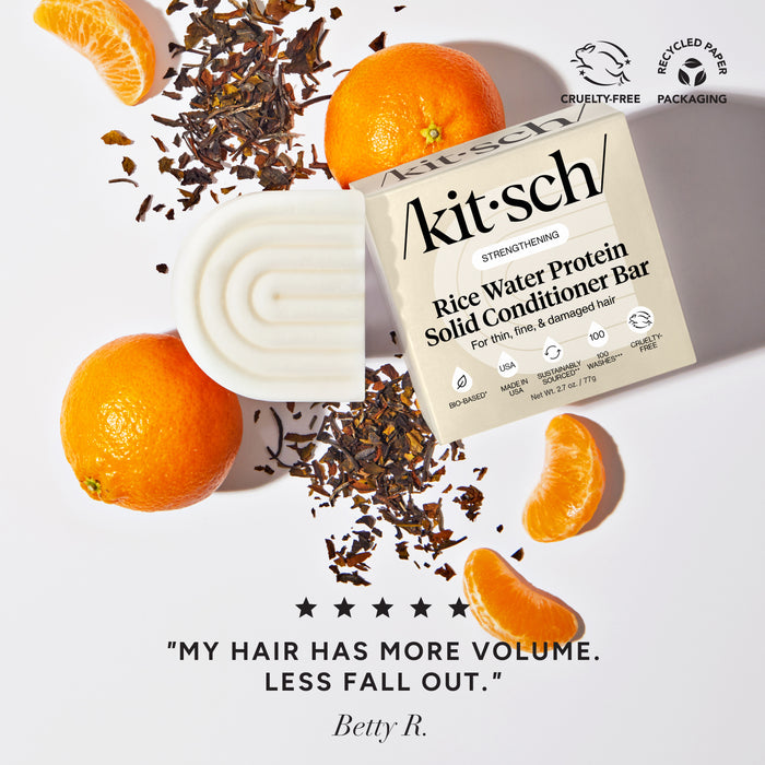 Kit'sch Rice Water Protein Solid Conditioner Bar with oranges and tea leaves on a plain background. There is a Leaping Bunny Cruelty-free badge and a recycled paper packaging badge in the upper right corner with a customer review at the bottom that reads "my hair has more volume, less fall out"