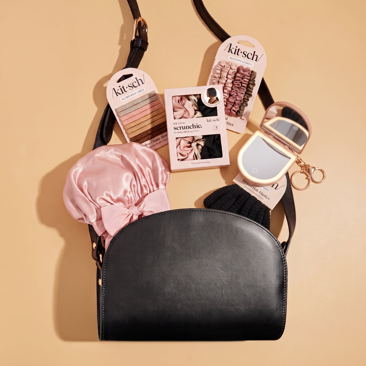 A black faux-leather crossbody bag with a curved front, displayed on a warm beige background with various beauty accessories arranged around it, including pink satin hair bows, a compact mirror, a black knit hair scrunchie, and two Kitsch hair extension kits.
